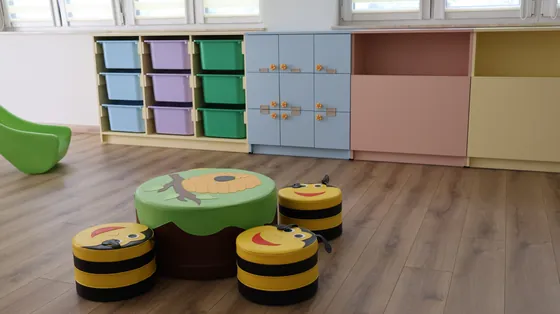 Sala żłobka z pastelową zabudową, regałem na pojemniki i szafkami pod oknami