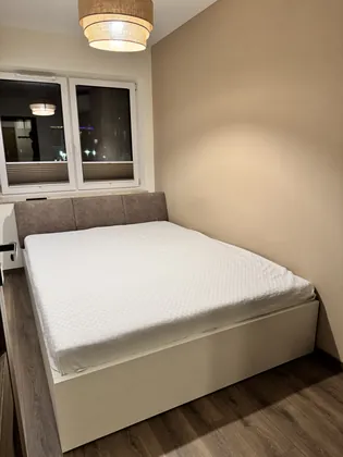 Łóżko z jasną ramą i tapicerowanym zagłówkiem pod oknem w sypialni.