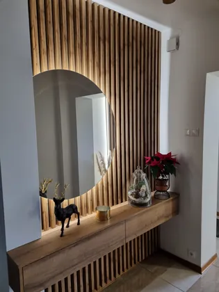 Przedpokój z drewnianymi lamelami, okrągłym lustrem i wiszącą konsolą z szufladami.